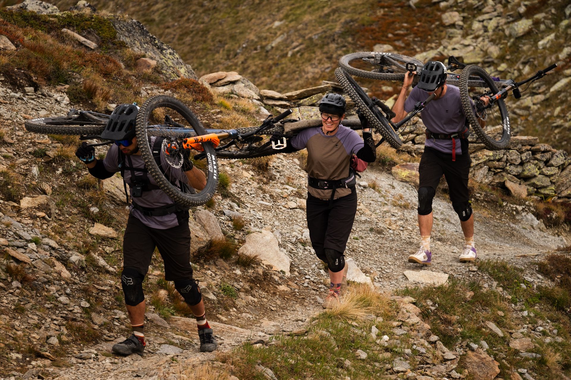 Three riders carrying bikes up a rocky trail